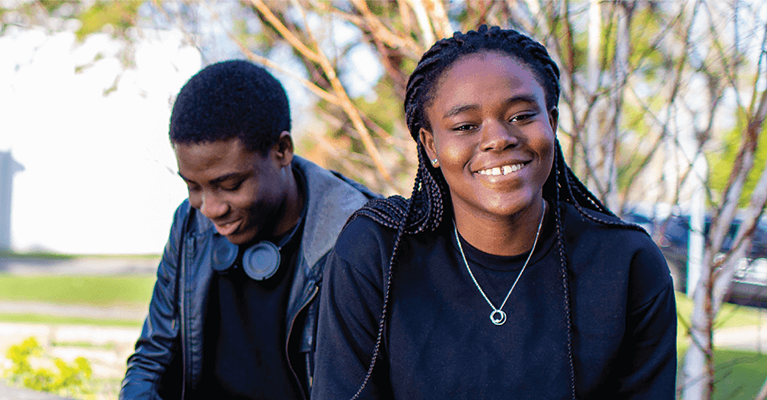 Two students sit under a tree on campus. The female student smiles at the camera while the male student is on his phone.